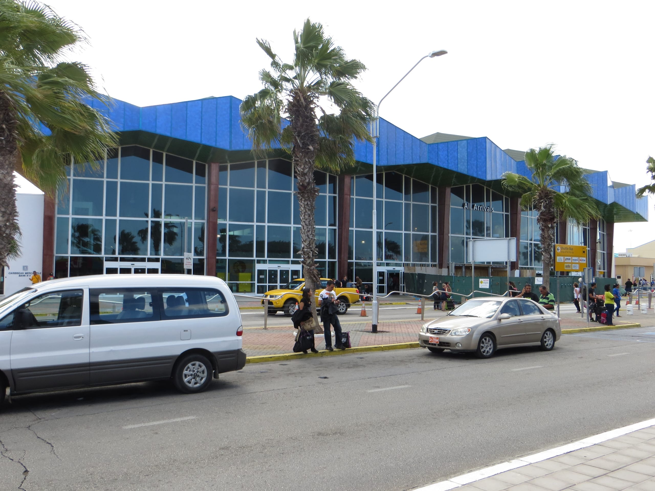 The glass front of the All Arrivals building at Queen Beatrix International Airport, Aruba, with palms and cars at the curb