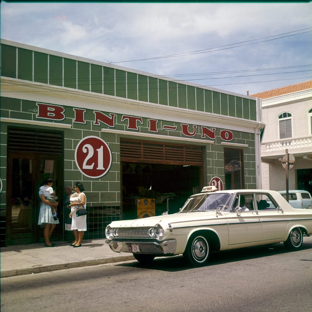 A cream 1960s taxi with a roof sign parked outside the Binti-Uno shop on Nassaustraat, Oranjestad, Aruba, in 1964