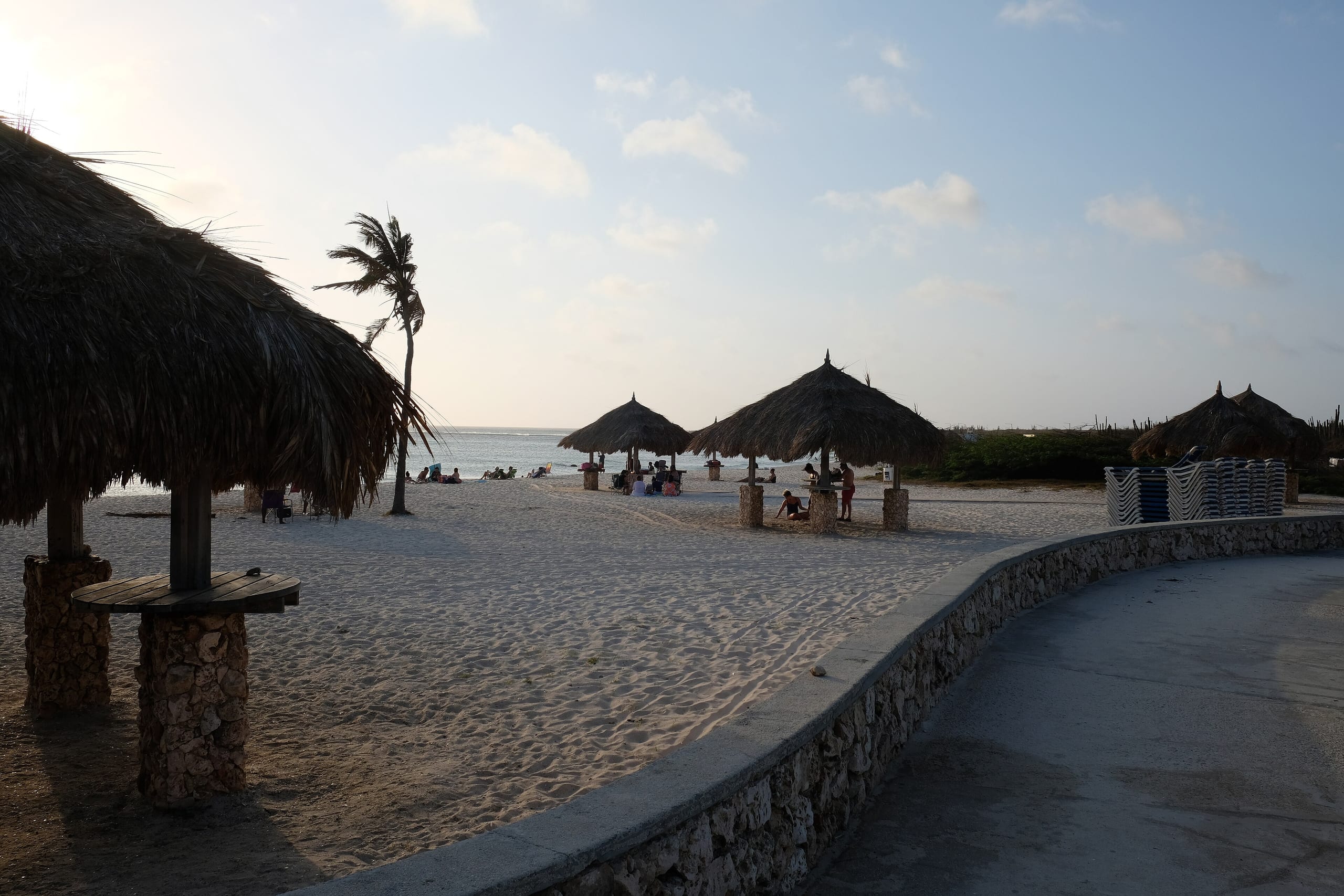 Thatched palapas on the sand at Arashi Beach, Aruba, behind a curved stone seawall in late afternoon light