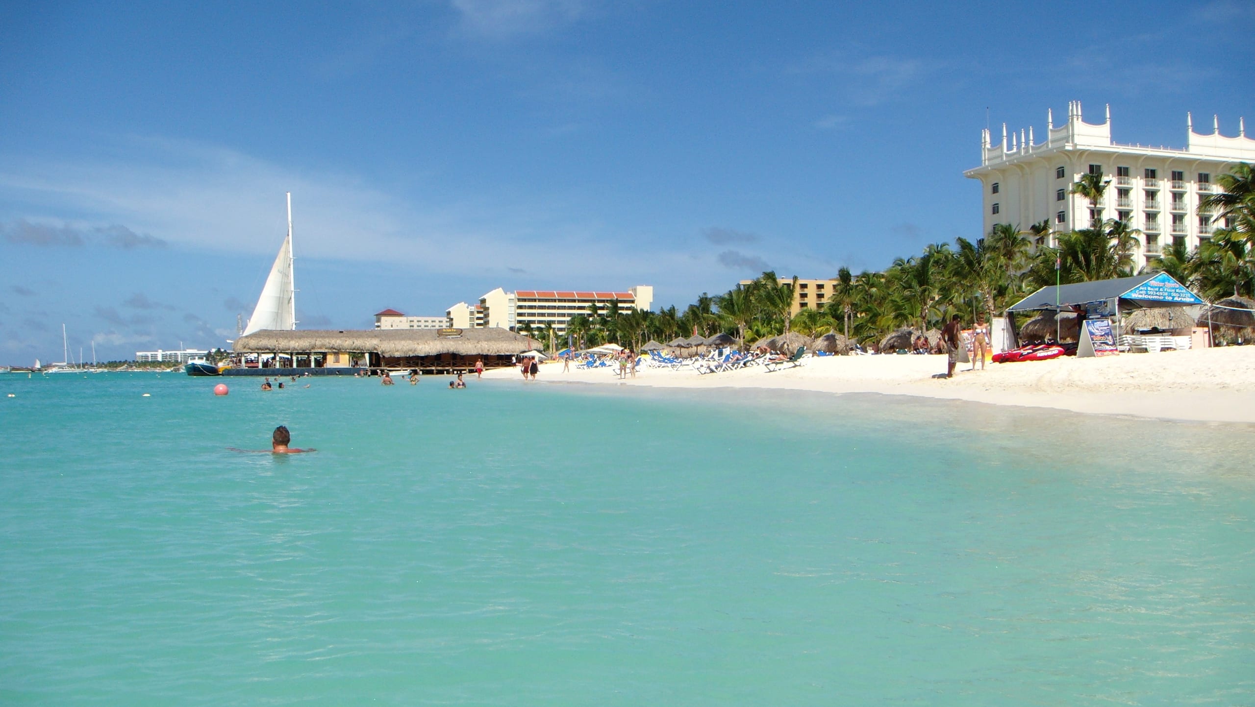 Swimmers in turquoise water off Palm Beach, Aruba, with a sailboat at a thatched pier, palapas on the sand and white hotel towers behind