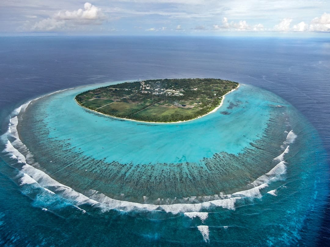 The island of Thoddoo in the Maldives ringed by its reef seen from above, surf breaking around the reef edge and shallow water inside it