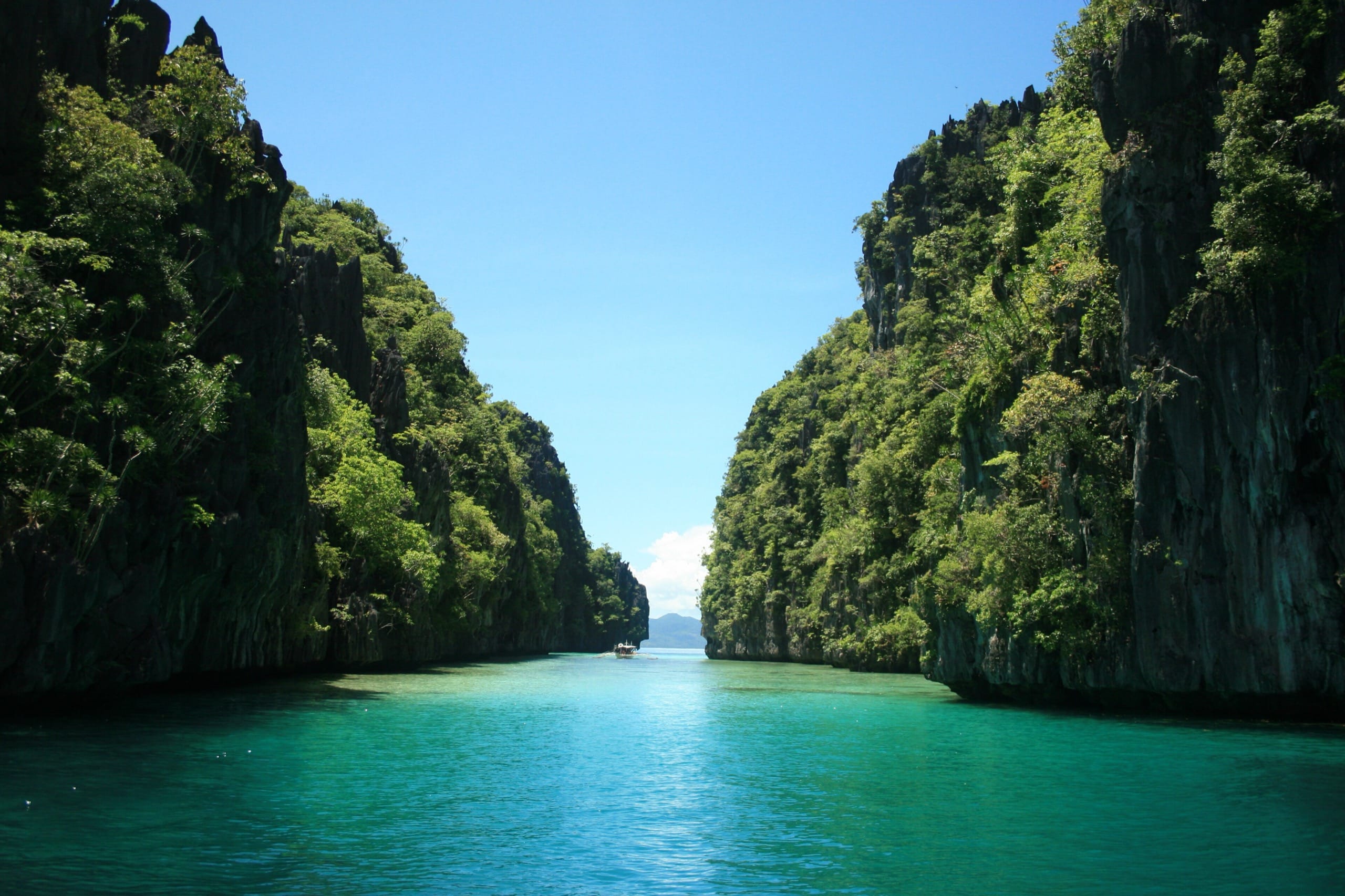 Narrow turquoise channel between steep forested limestone walls with a single outrigger boat far ahead