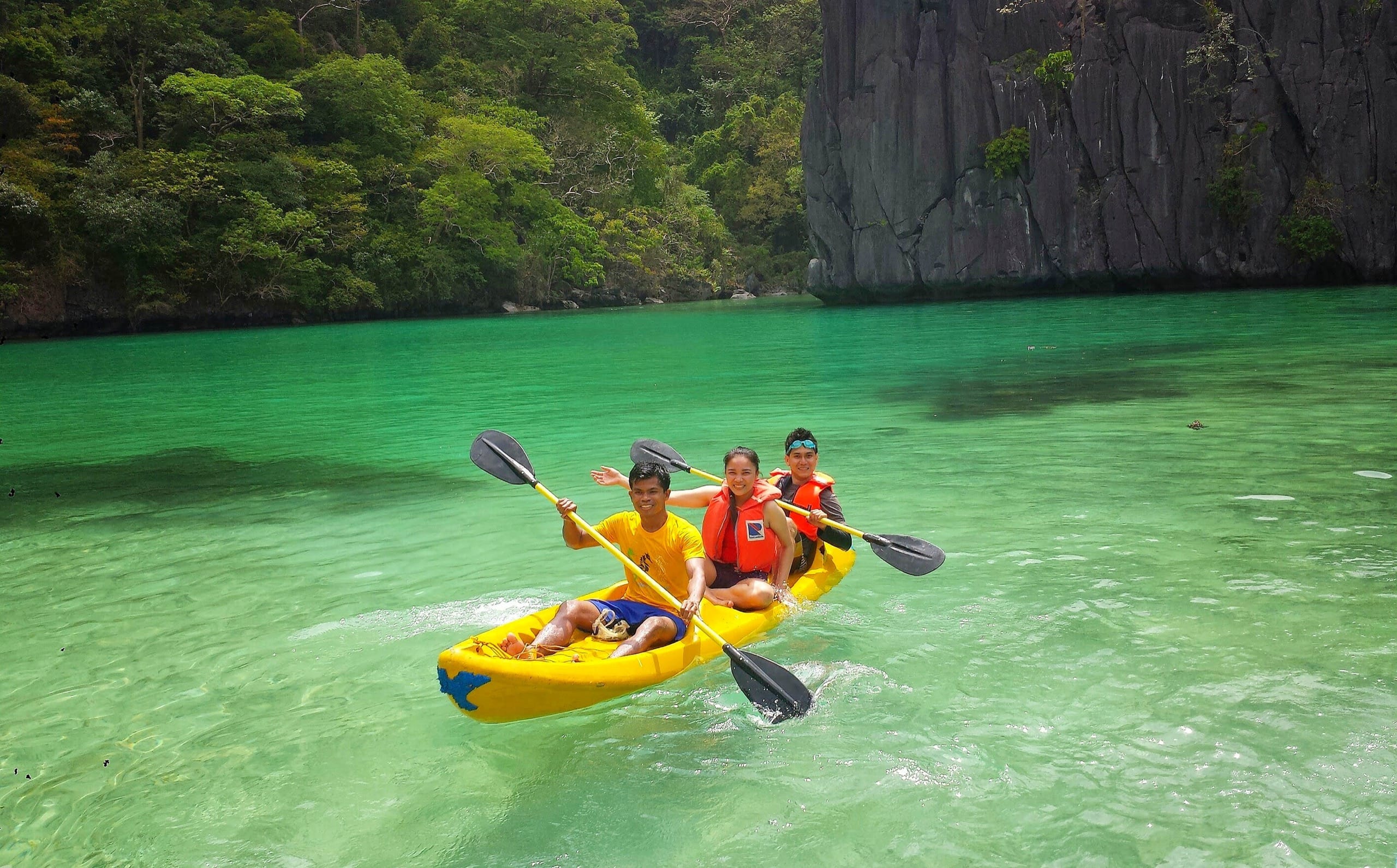 Three people paddling a yellow tandem kayak on bright green lagoon water beside a dark limestone wall