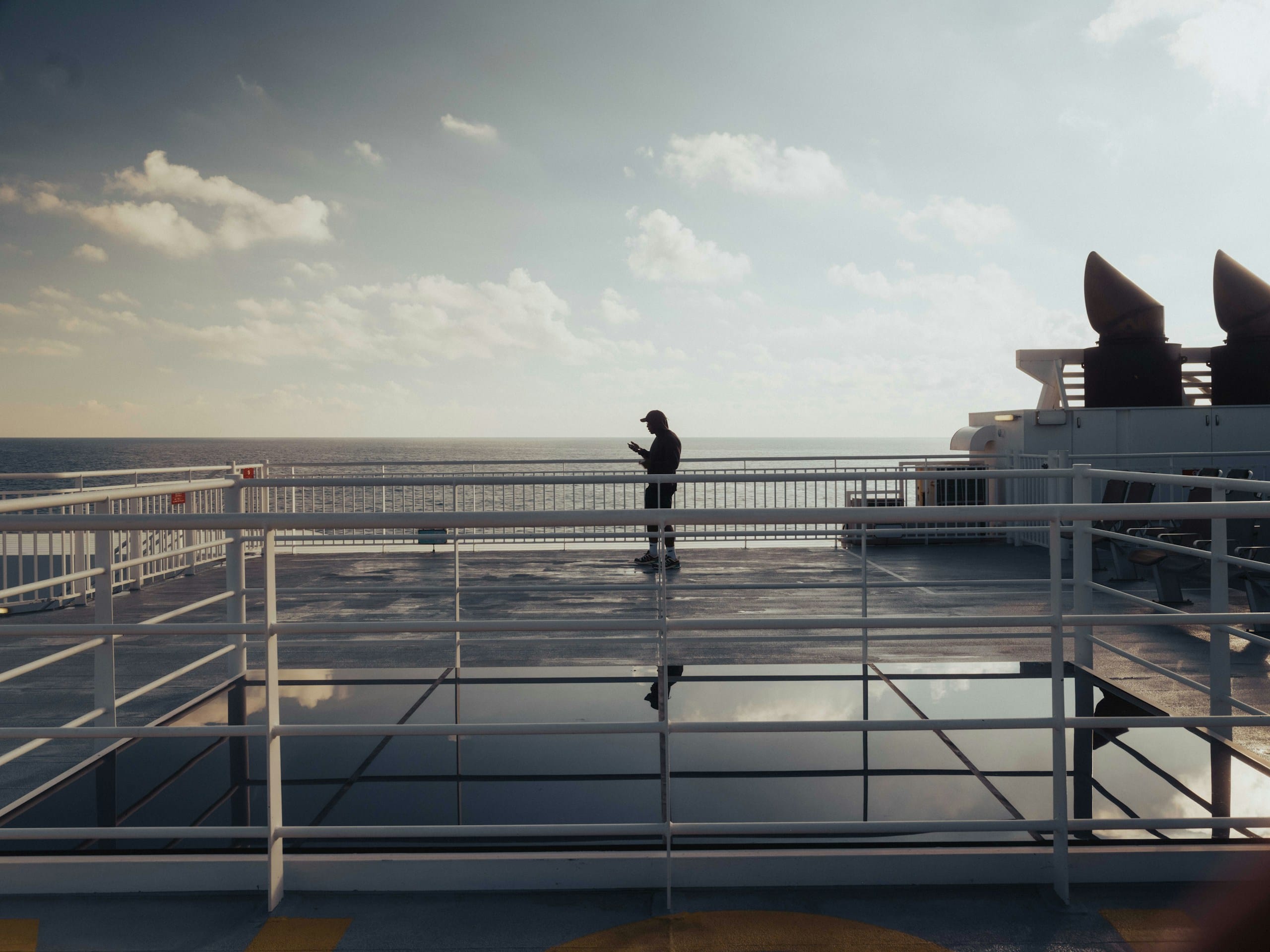 A lone passenger reads a phone on the open deck of a ferry, flat sea and an evening sky beyond the railings