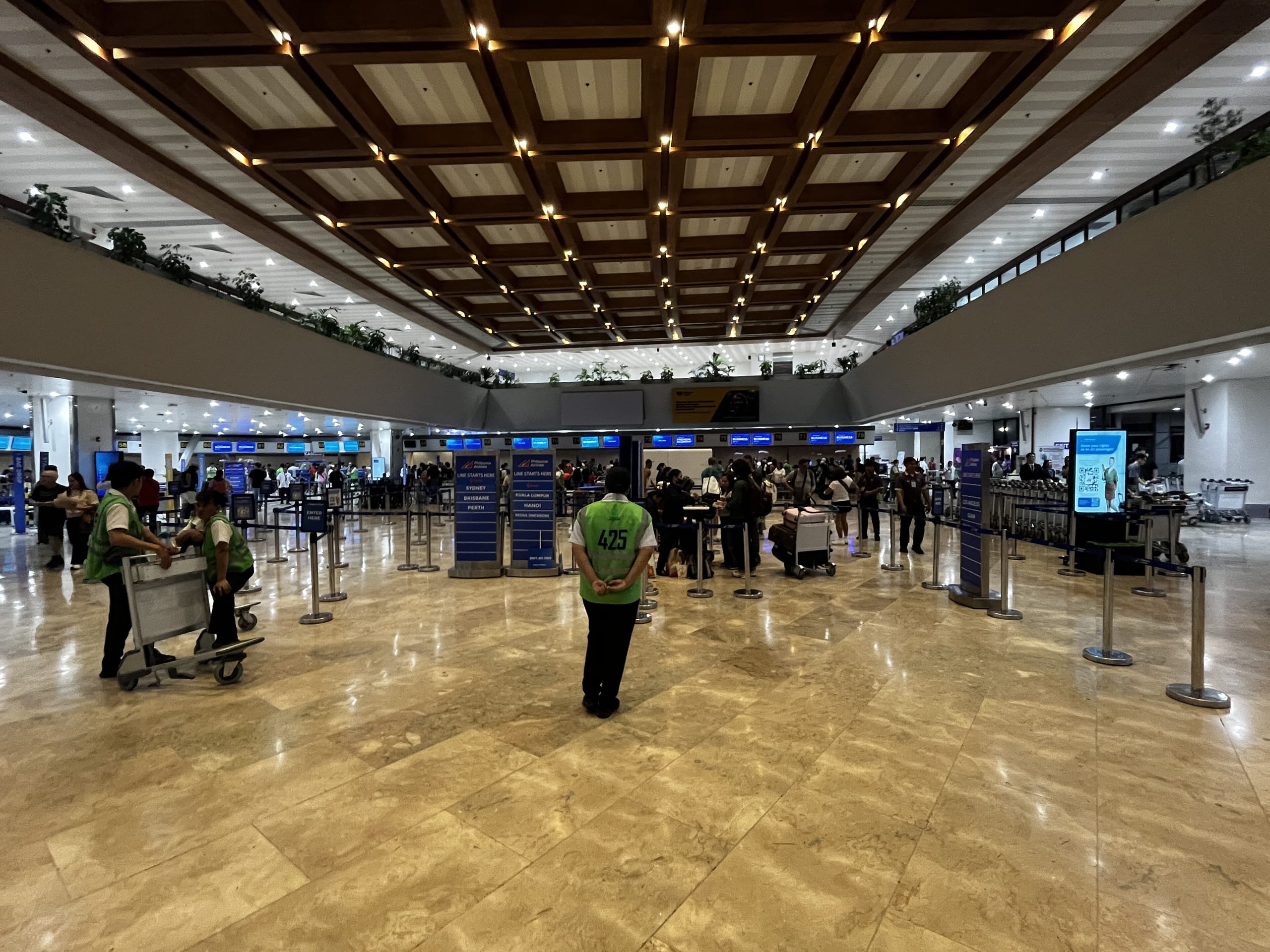 Check-in hall at Manila Ninoy Aquino Terminal 1, queues under a coffered wooden ceiling and an attendant in a green vest