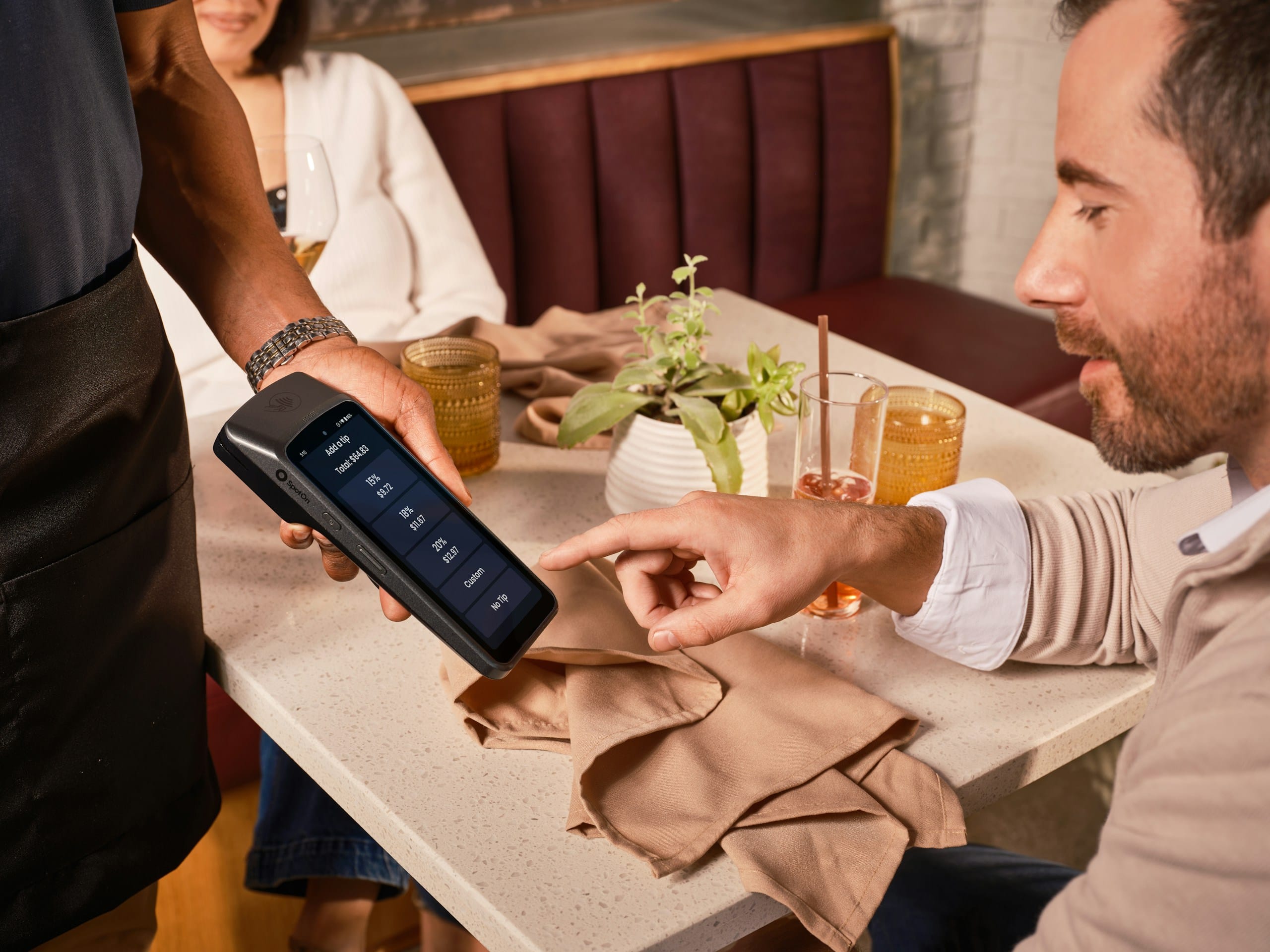A server holding a card reader with tip percentages while a diner taps one at a restaurant table