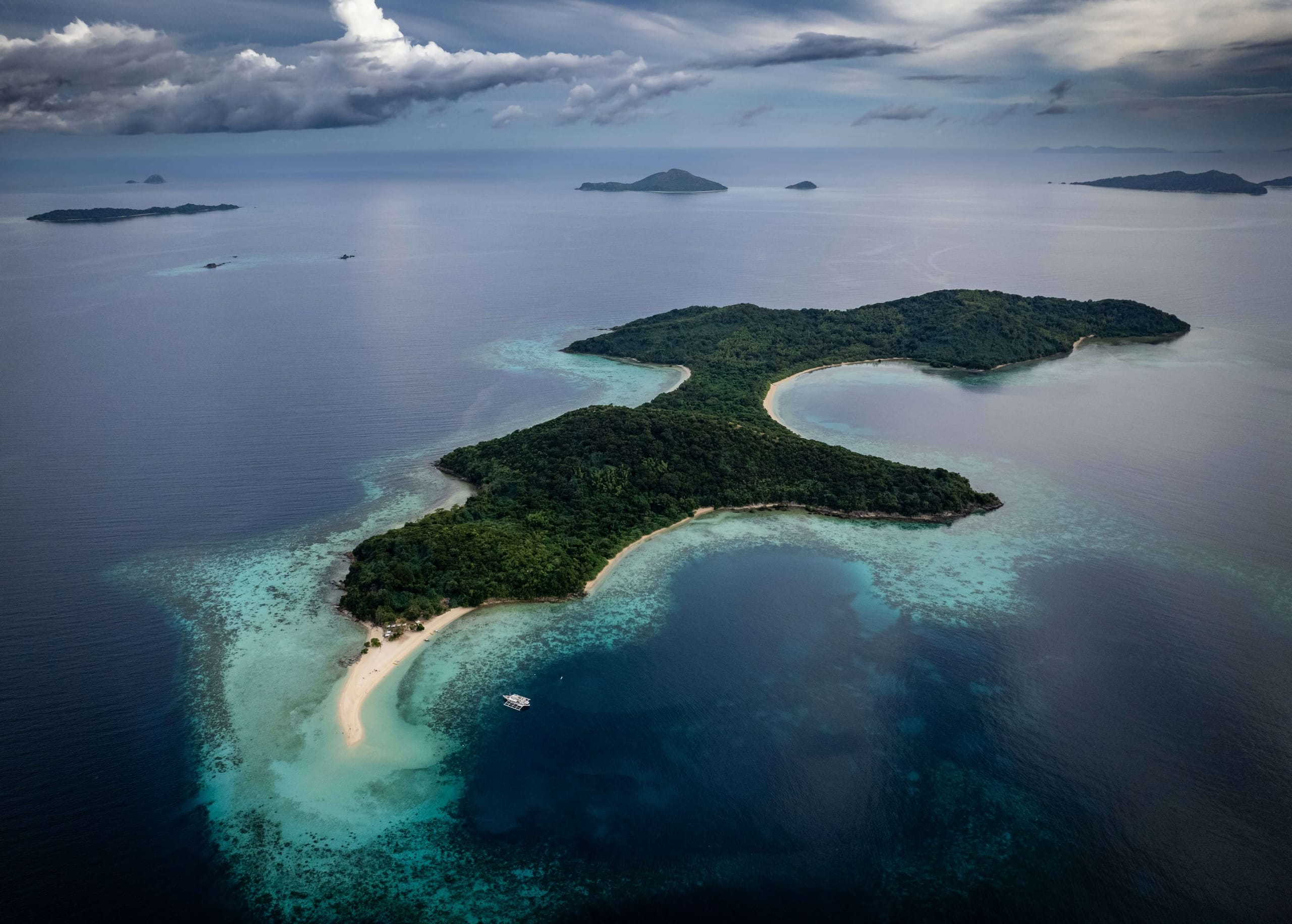 A low green island lying in pale shallows seen from above, a sandbar running off one end and deeper blue water around it