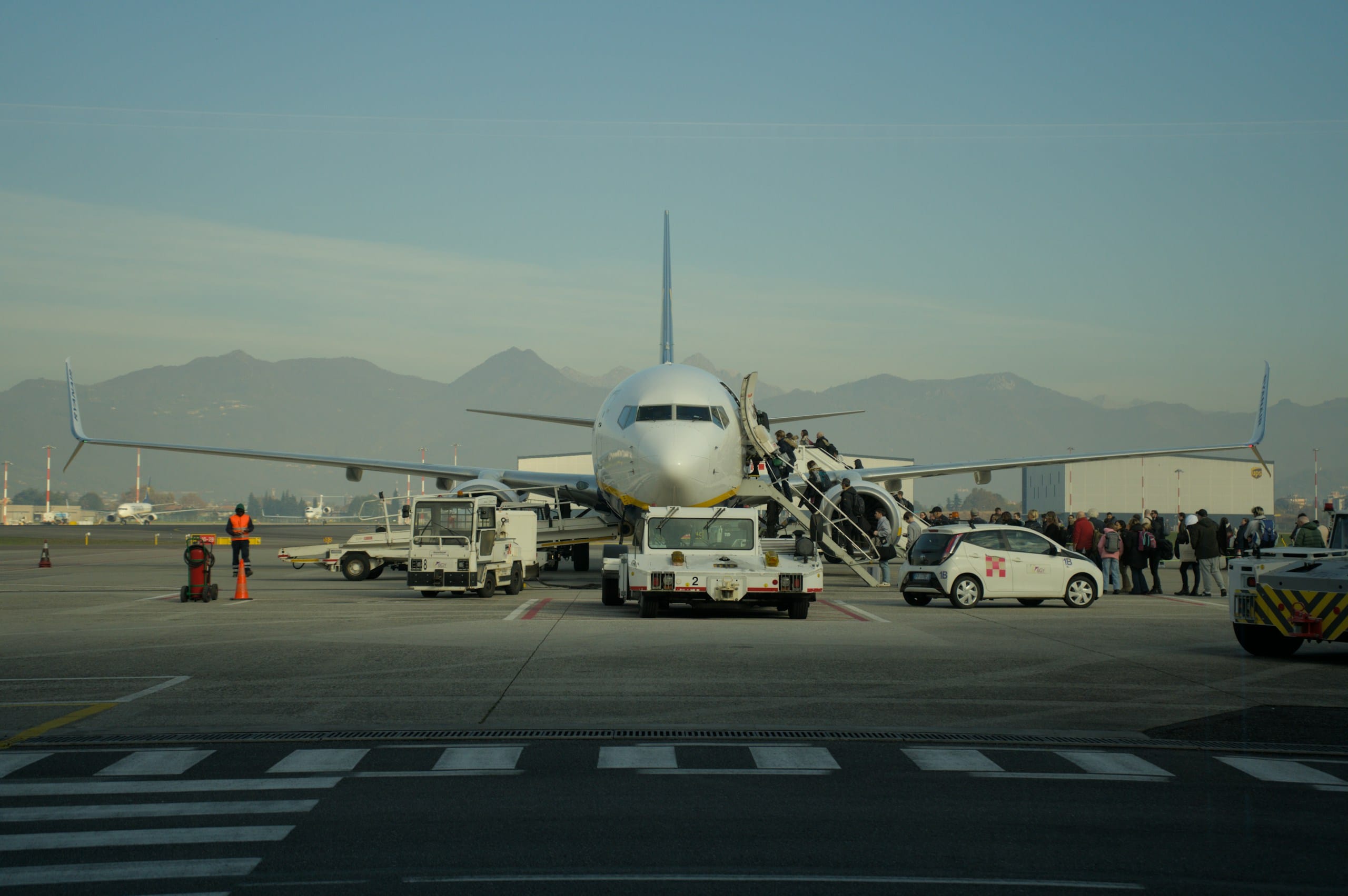 Passengers climbing the boarding stairs to a jet parked on a tarmac, mountains hazy behind
