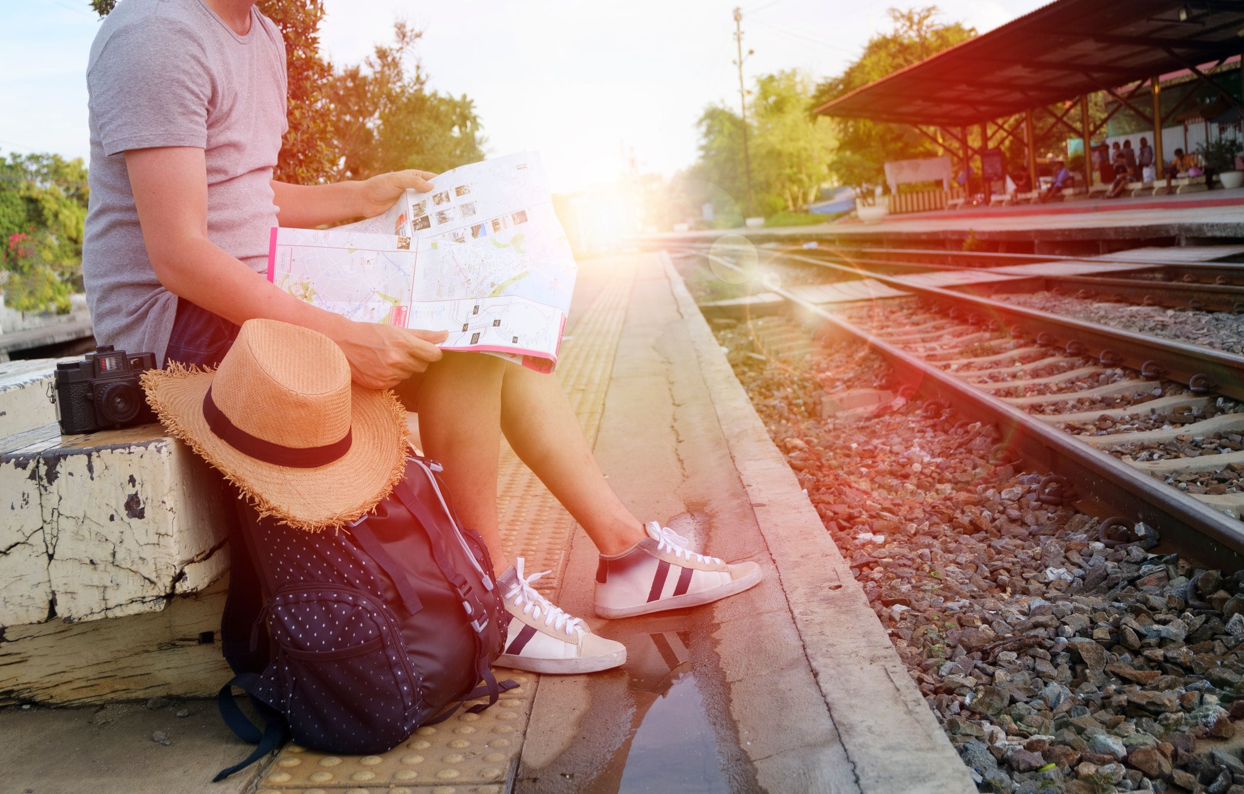A traveler on a station platform reading a folded paper map, a backpack and straw hat at their feet
