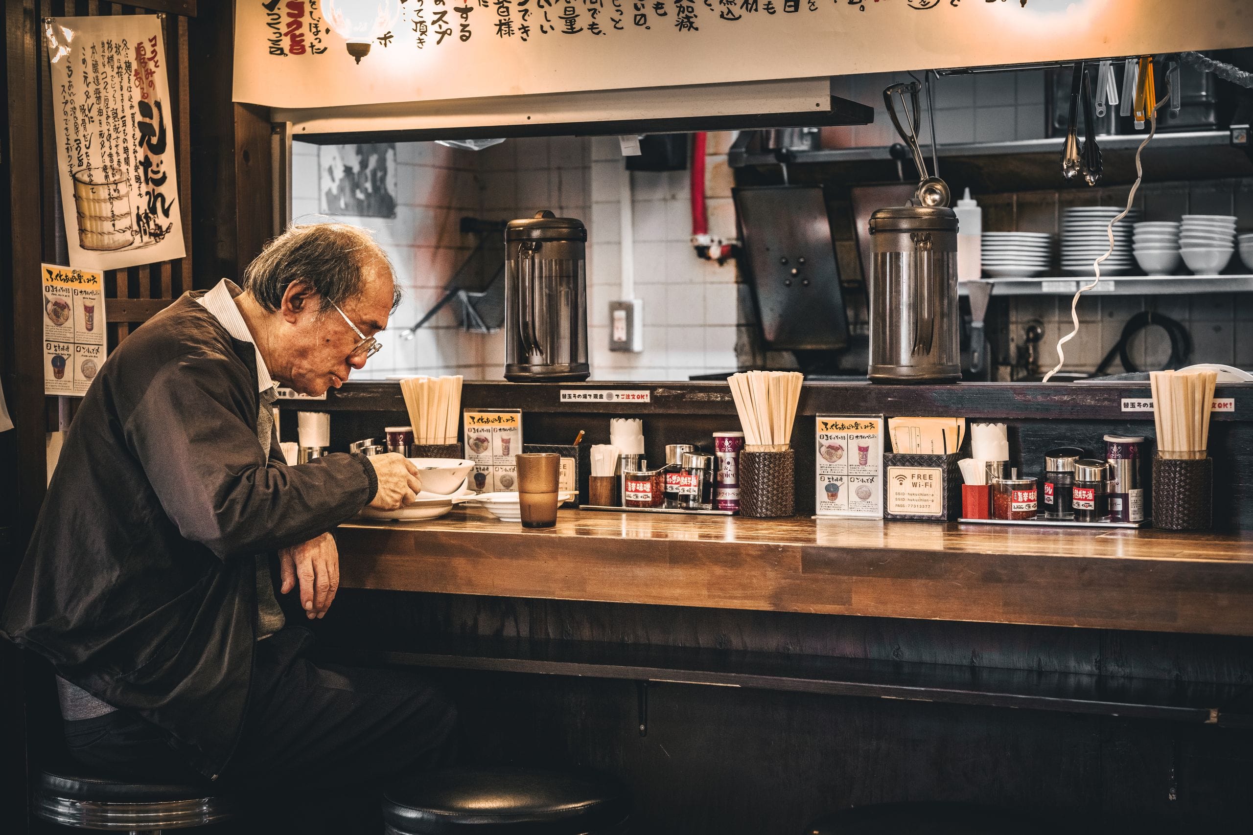 An older man eating alone at the counter of a small ramen shop in Tokyo, chopsticks and condiments along the bar