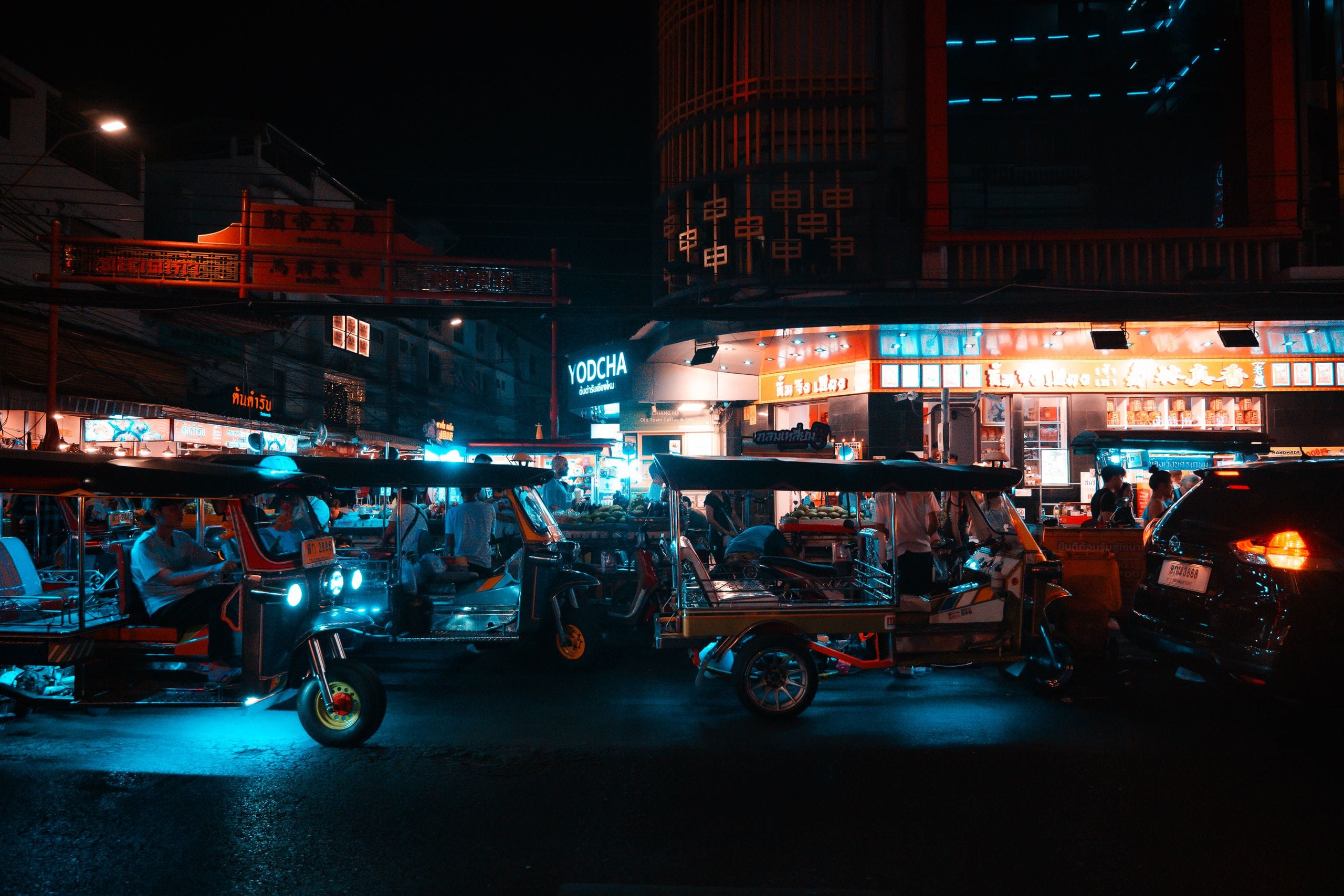 Tuk-tuks lined up at night under neon shop signs on a Bangkok street corner