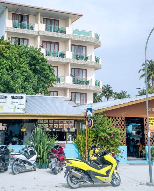 A four-story guesthouse with balconies above two small shopfronts on a street in Maafushi, scooters parked at the front and a coffee board hanging by the door.