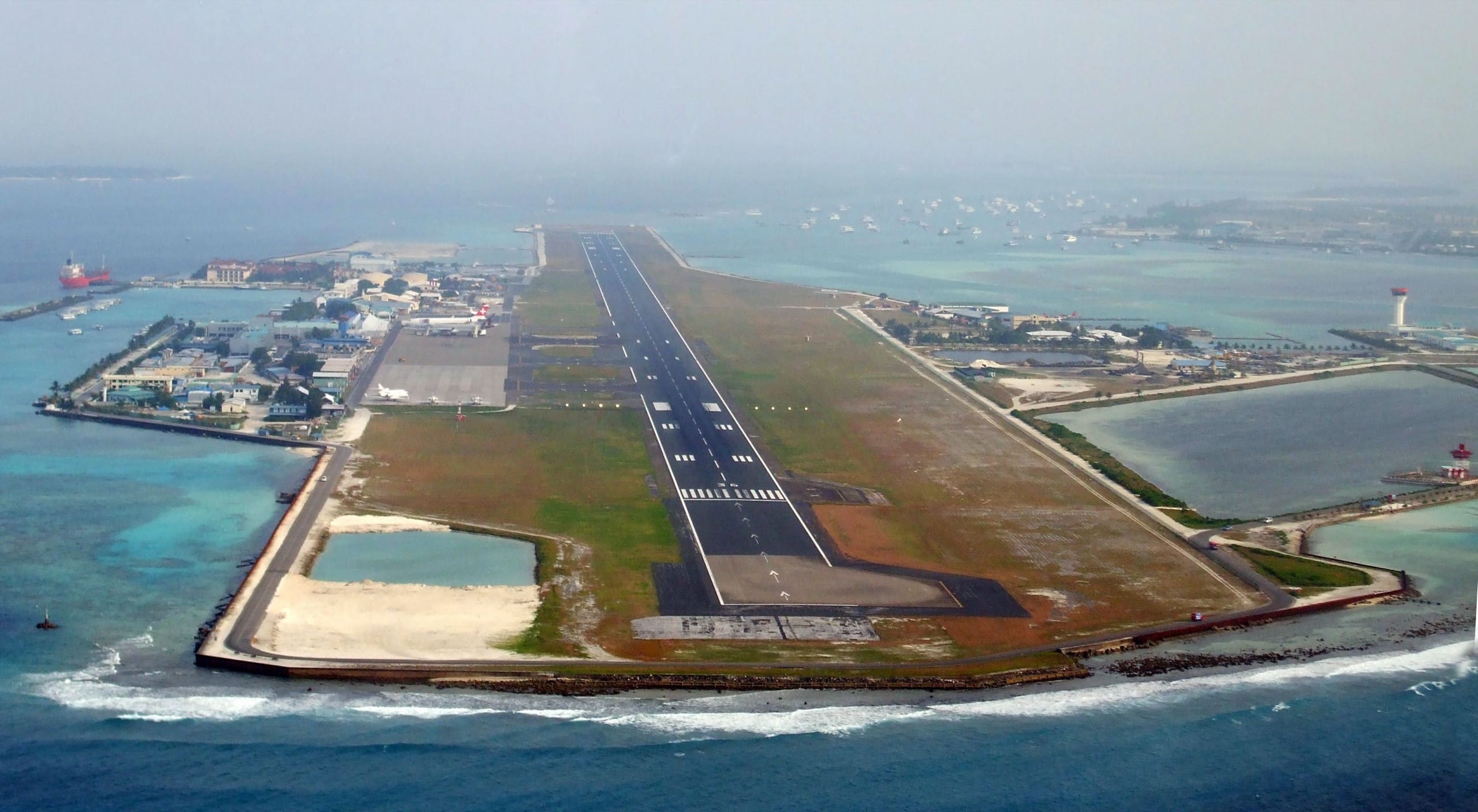 Aerial view of the Velana airport island, a single runway on reclaimed land ringed by reef and sea, with Malé beyond
