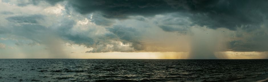 A dark cloud bank over open sea with rain falling from it in gray columns, and a strip of pale light along the horizon