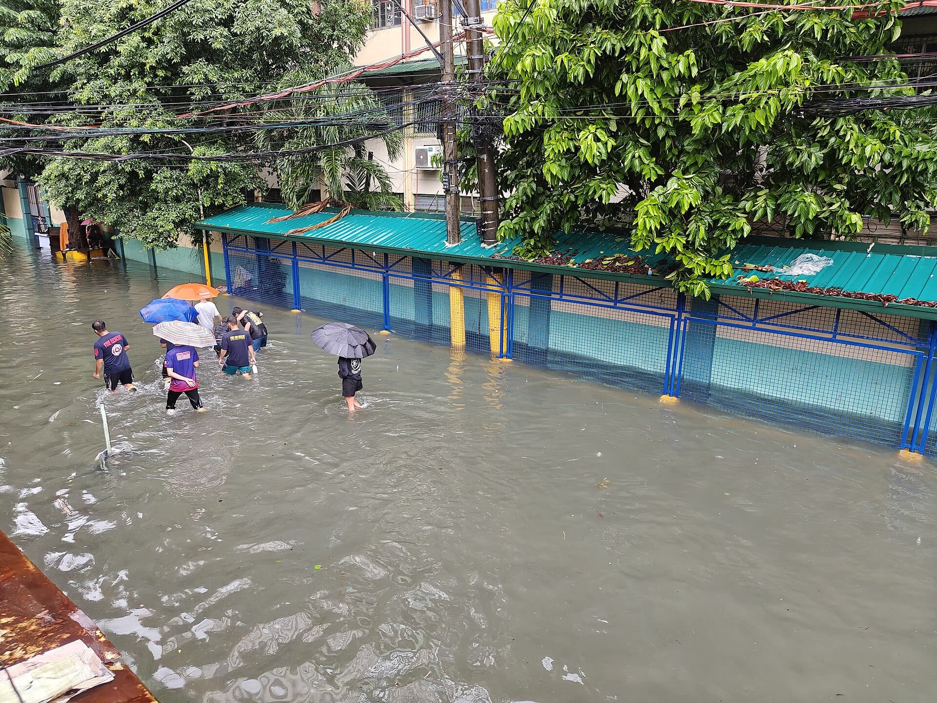 People with umbrellas and rain gear wading through knee-deep floodwater on a street in Quiapo, Manila