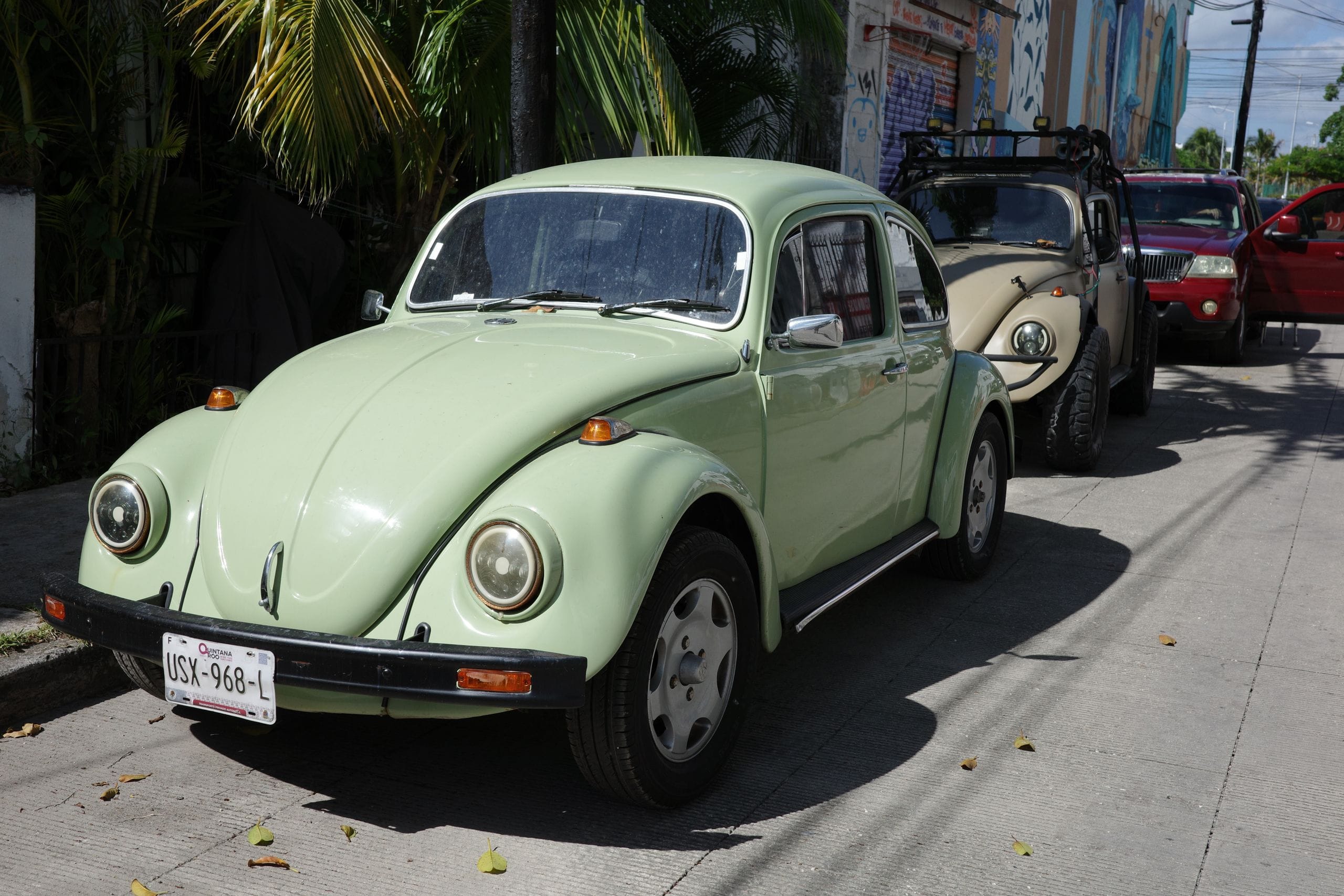 Pale green Volkswagen Beetle with Quintana Roo plates parked on a shaded street beside a mural wall