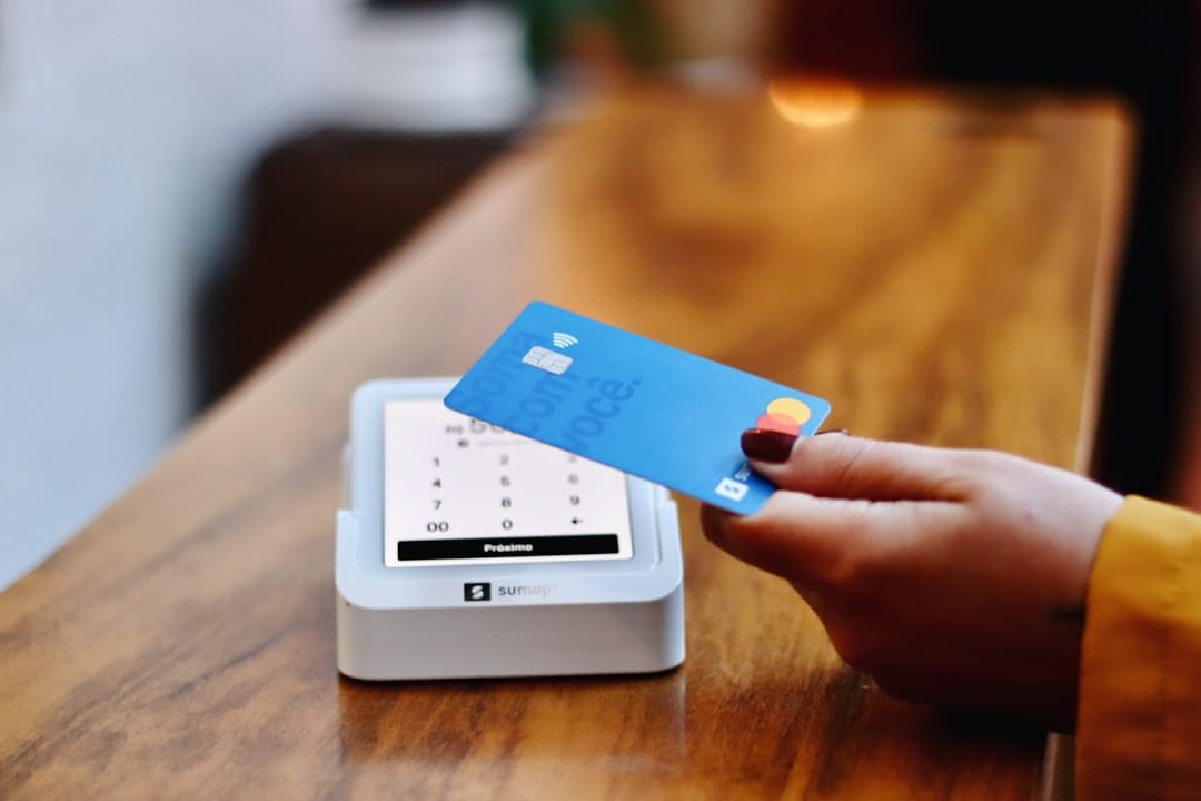 A hand holding a blue contactless card up to a white card reader standing on a wooden counter