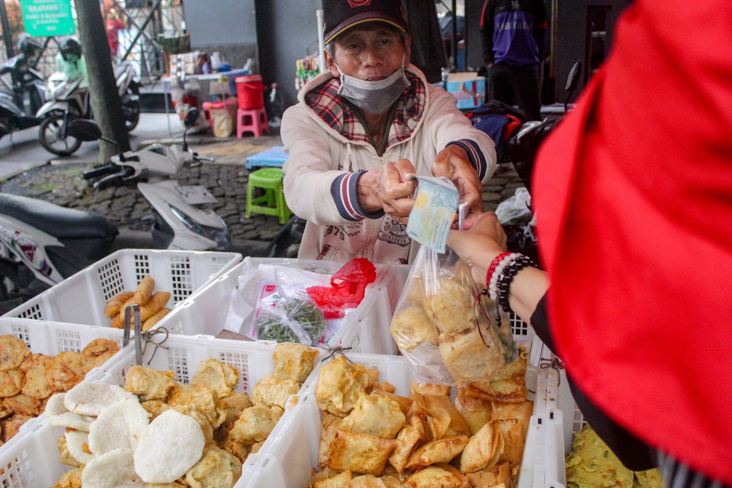 A vendor in a face mask passing a bag of fried snacks to a customer holding a blue banknote
