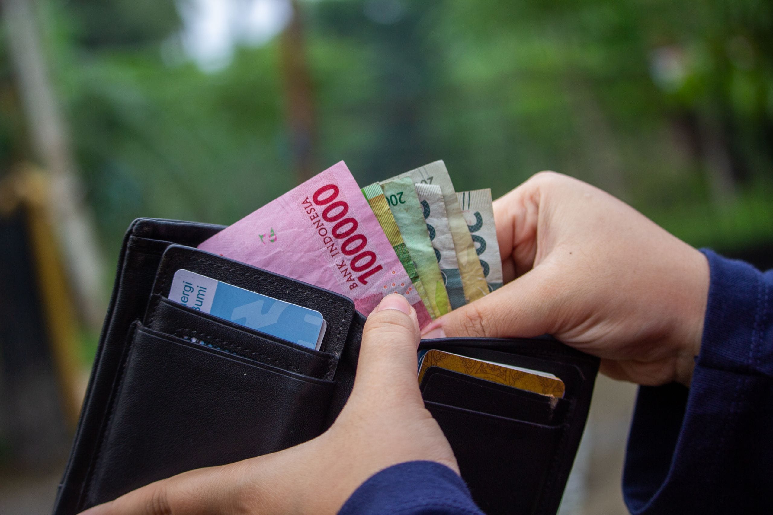 Two hands opening a black wallet holding a blue card and a fan of Indonesian banknotes