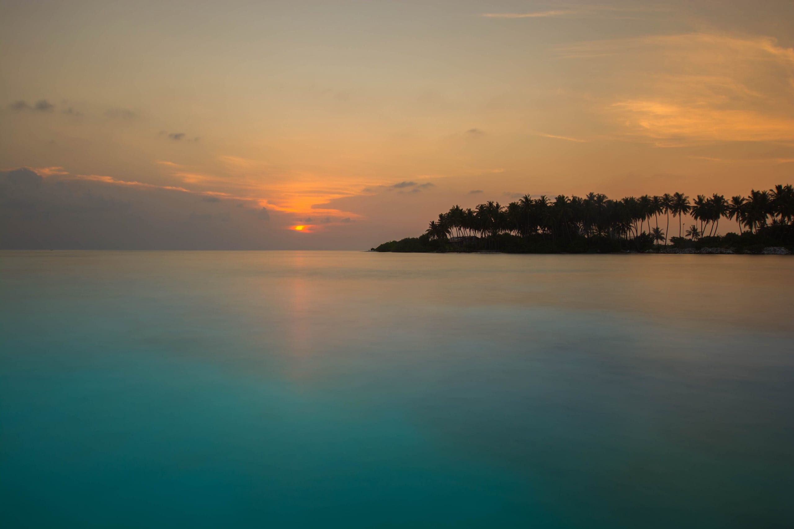 The sun setting over lagoon water with a palm-lined island shore in silhouette