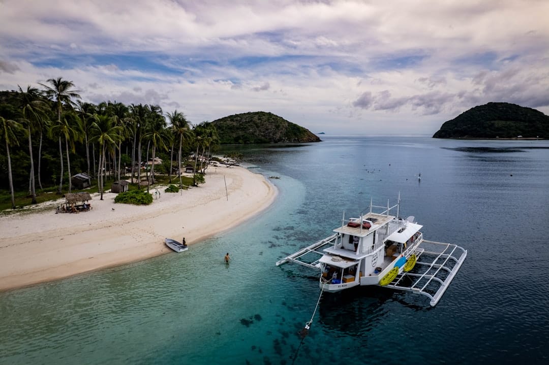 A white sand spit in the Philippines lined with coconut palms seen from above, a passenger outrigger boat moored offshore and two green islands on the horizon