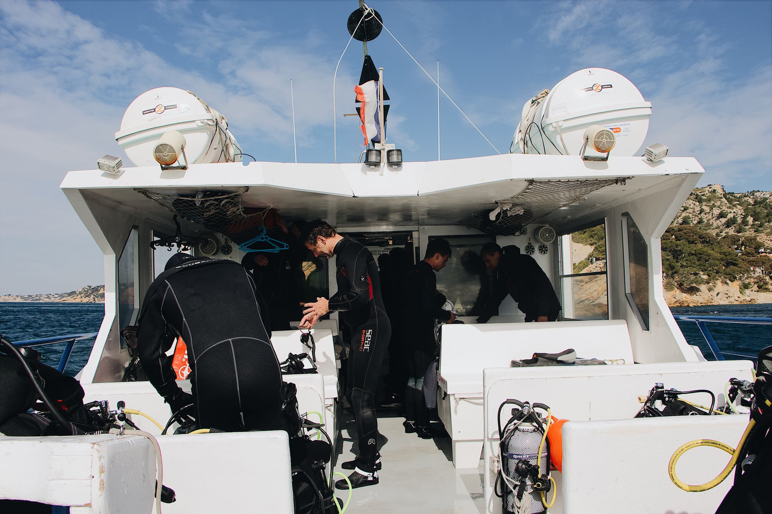 Divers in black wetsuits gearing up on the deck of a dive boat, tanks and regulators racked along the benches