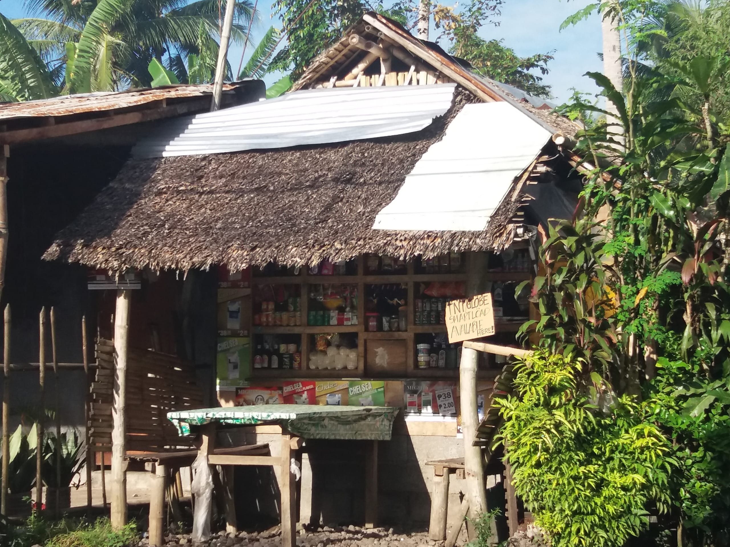 Hand-lettered cardboard sign reading TNT Globe Smart load available here beside shelves at a thatched roadside store