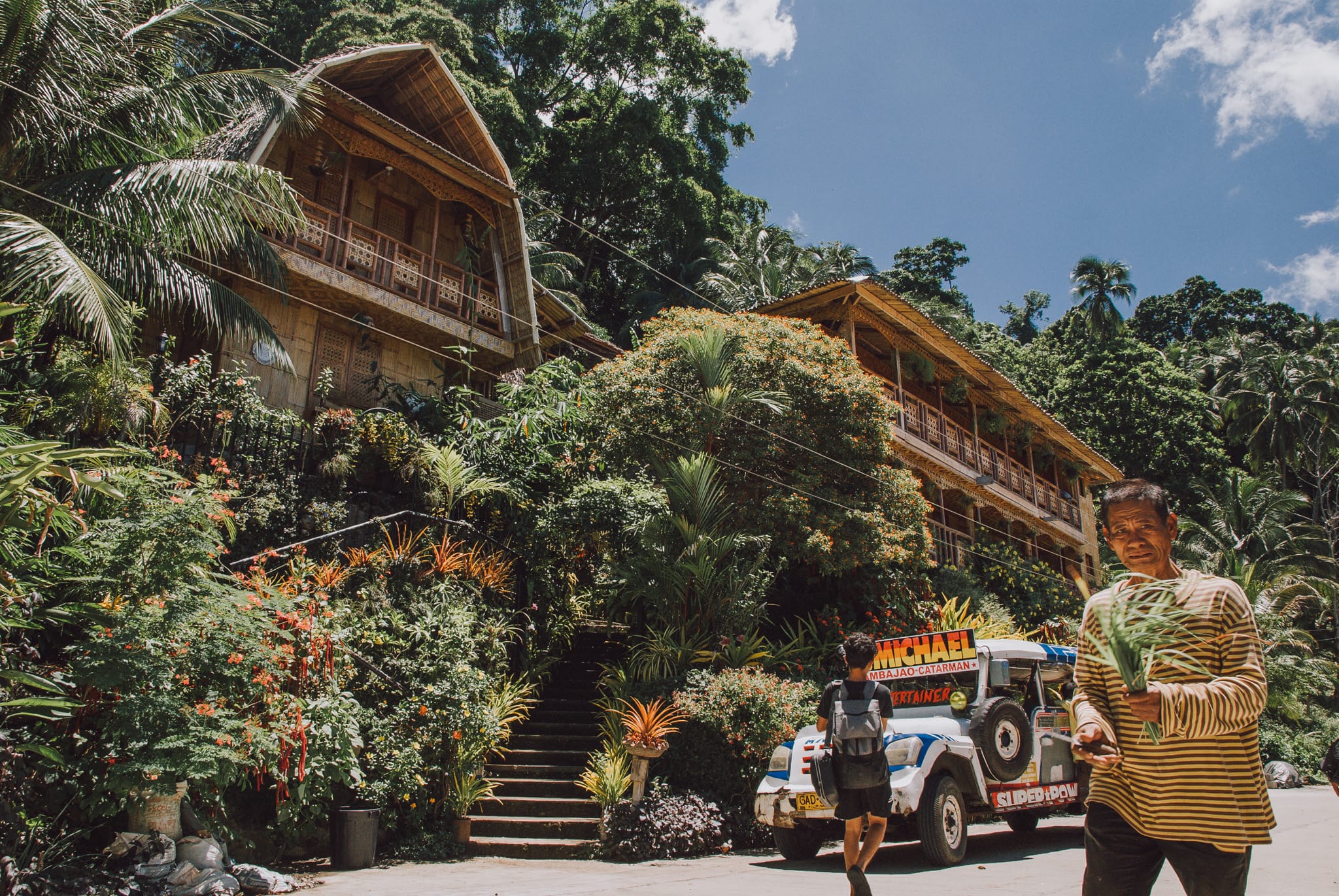 Wooden and bamboo building with carved balconies climbing a green hillside, a jeepney parked below, Camiguin