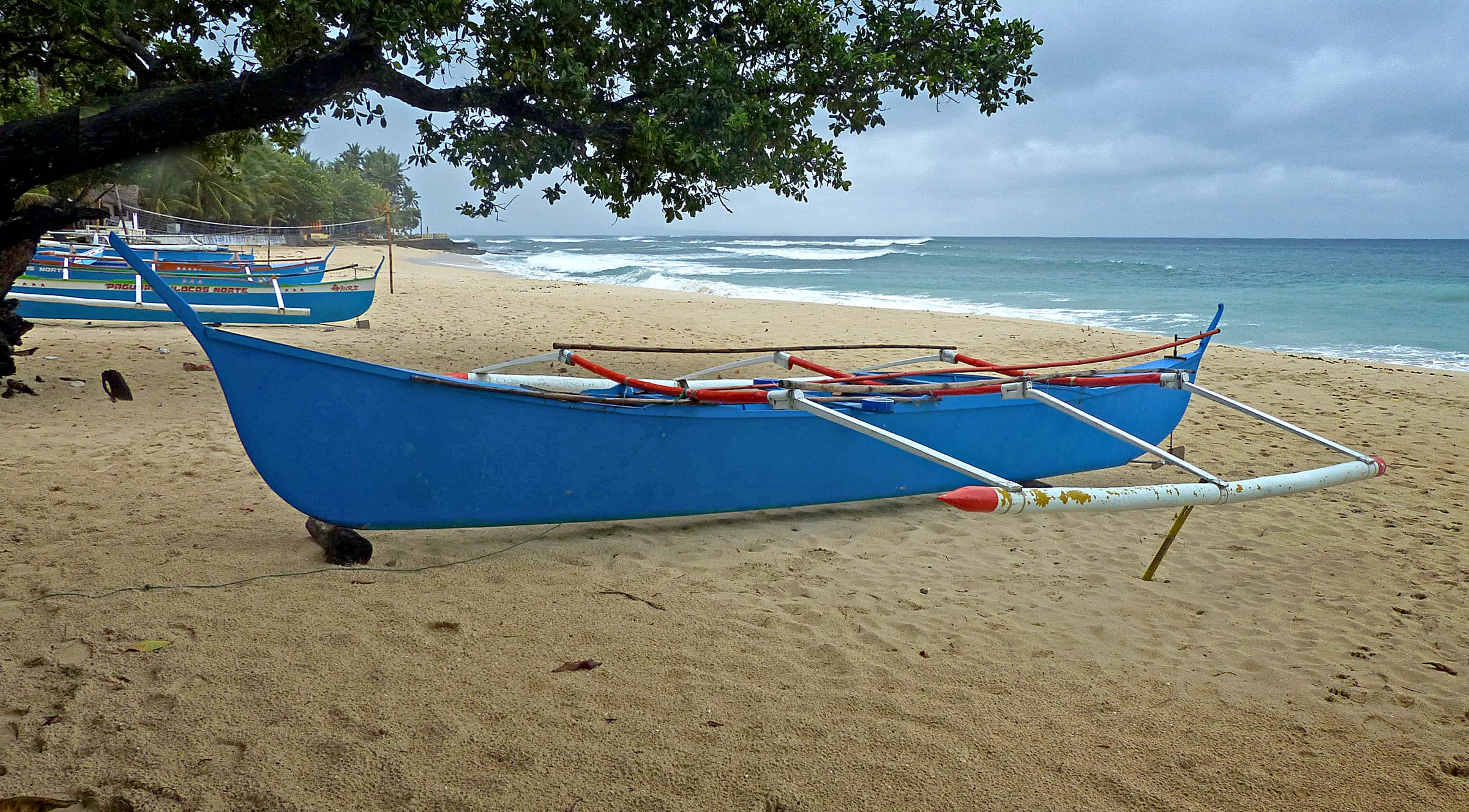 A blue outrigger bangka hauled up on a sandy beach, surf running in behind it and more boats along the shore