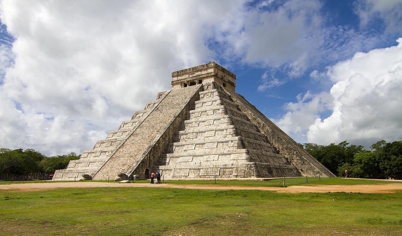 El Castillo at Chichen Itza seen corner-on across the grass plaza, a small group of visitors standing at the foot of the near stairway
