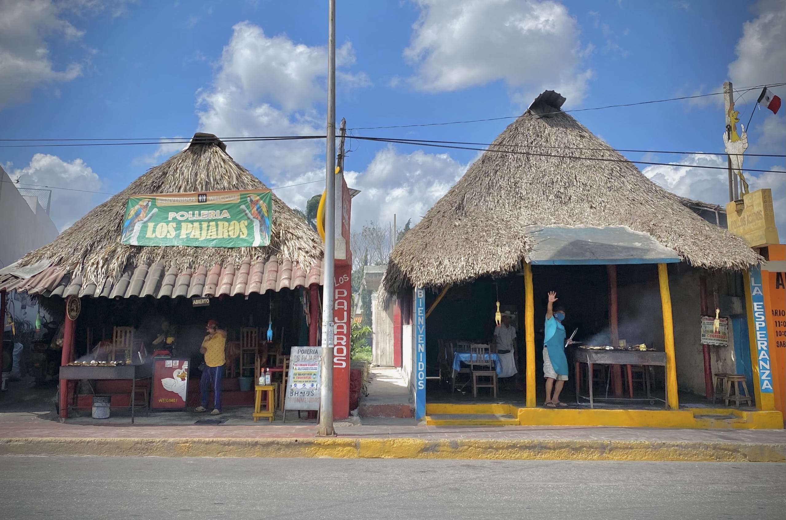 Two thatched roadside grill restaurants on a sunny street in Pisté, a cook waving from one