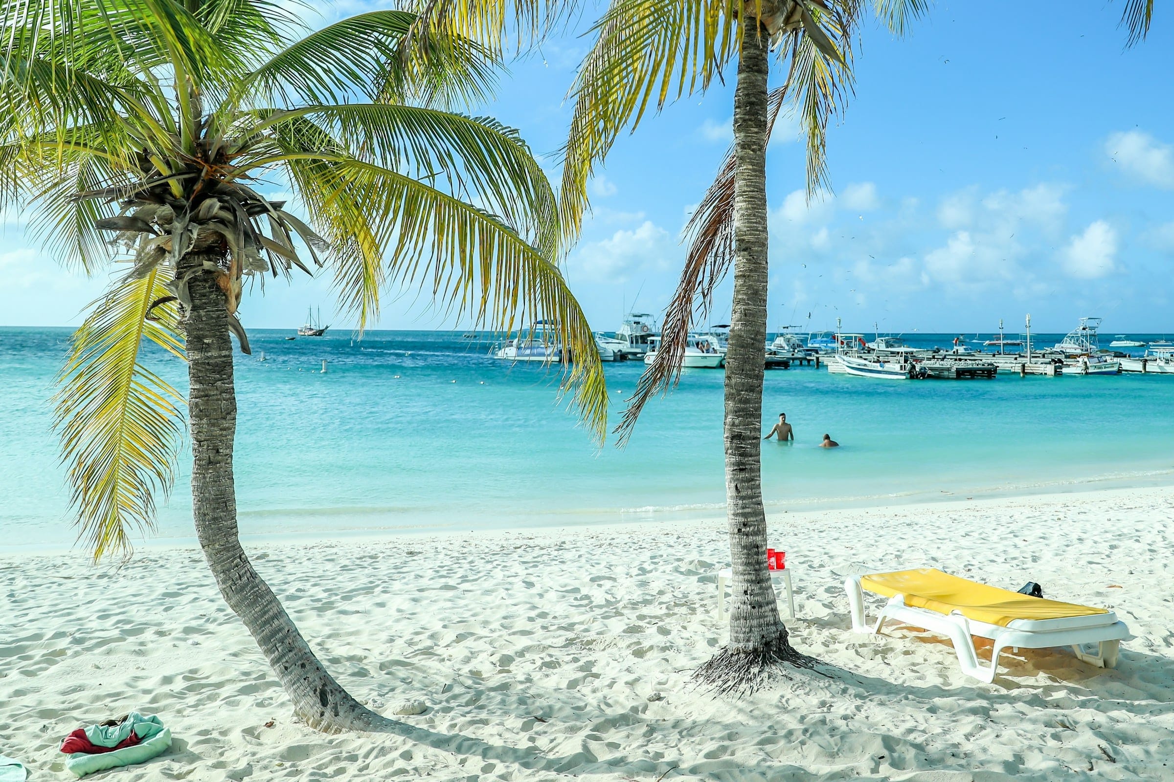2 palm trees leaning over white sand, a yellow lounger in their shade and boats moored out in the turquoise water