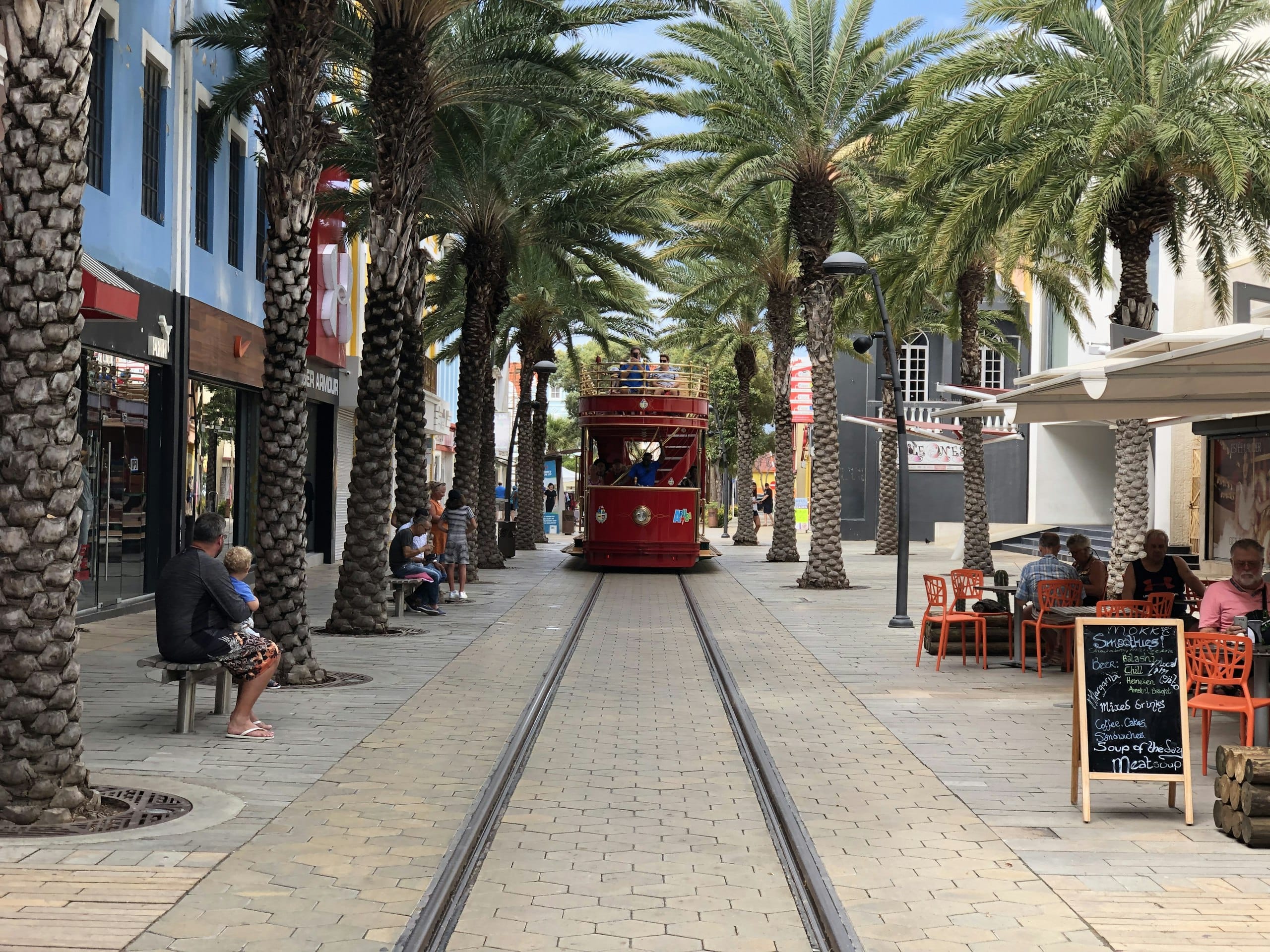 The red Oranjestad streetcar coming down Main Street between rows of palms, café tables along the side