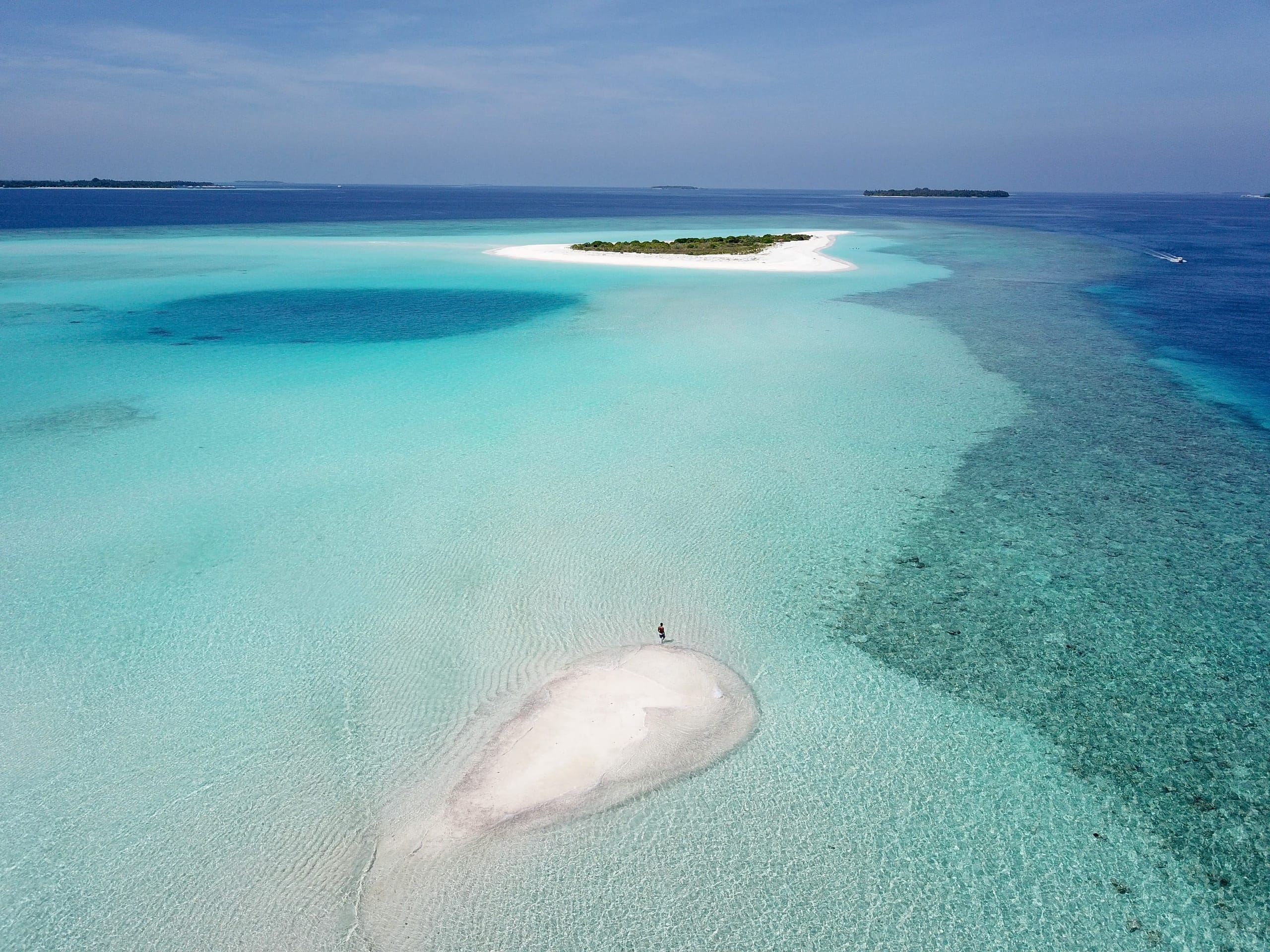 A pale sandbank in a wide turquoise lagoon with a single person standing on it