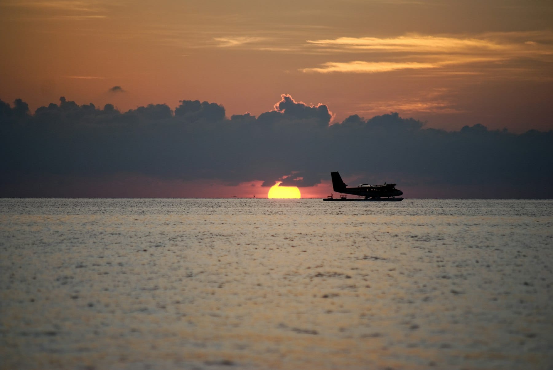 A seaplane sitting on flat open water with the sun dropping to the horizon behind it