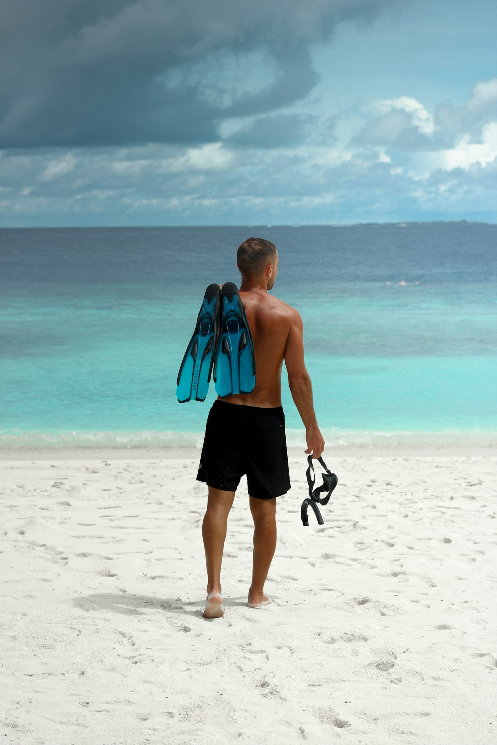 A man walking across white sand to the water carrying his own fins and mask