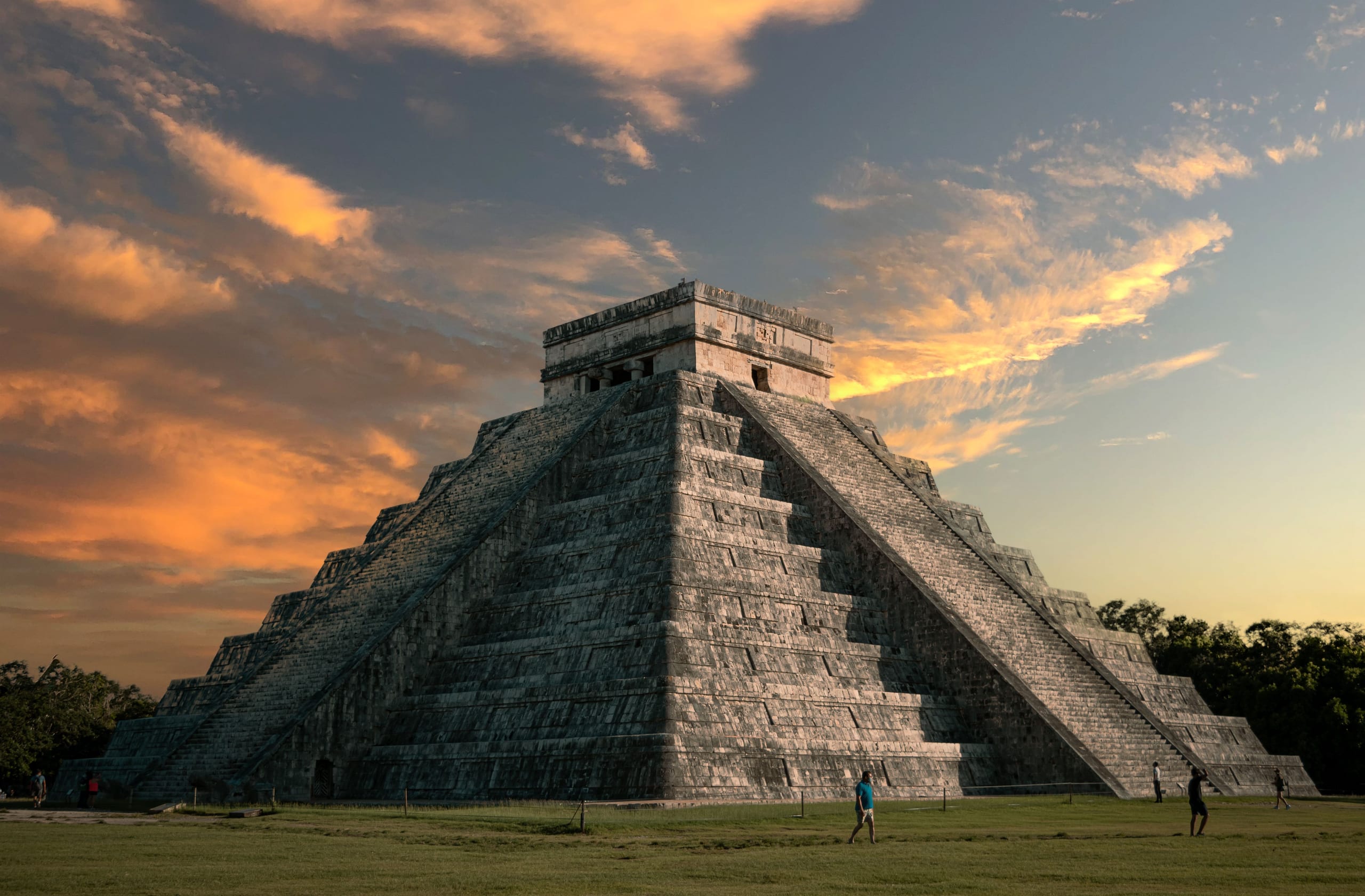A stepped stone pyramid on a mown lawn with the sky going orange behind it and a few people at its foot