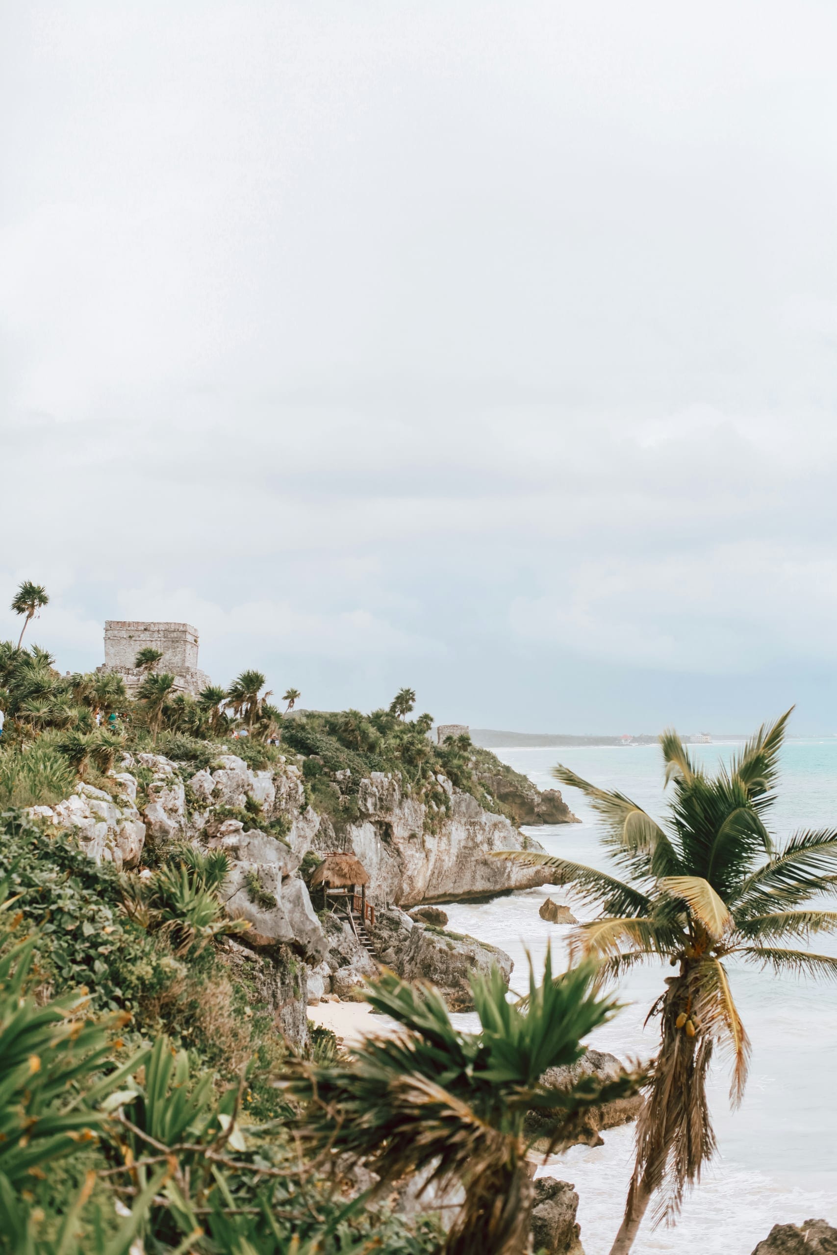 A low stone building on a rocky point with palms and scrub around it and the sea pale behind