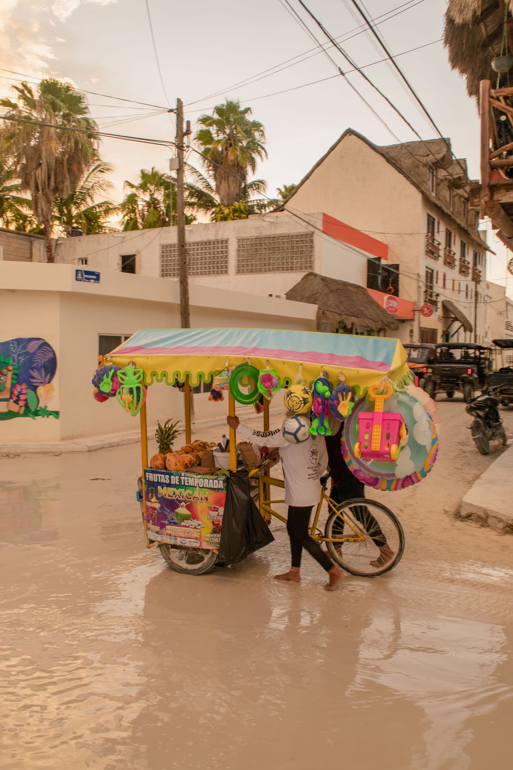 A fruit cart under a painted awning hung with cut fruit and papel picado, parked on a street corner